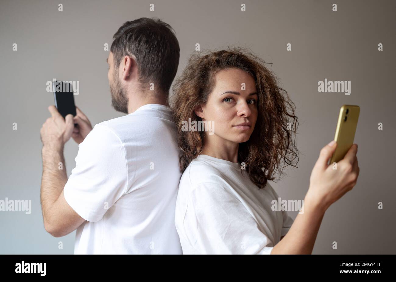 Frau und Mann, die weiße T-Shirts tragen, stehen Rücken an Rücken und benutzen ihre Handys getrennt, Frau schaut in die Kamera. Stockfoto