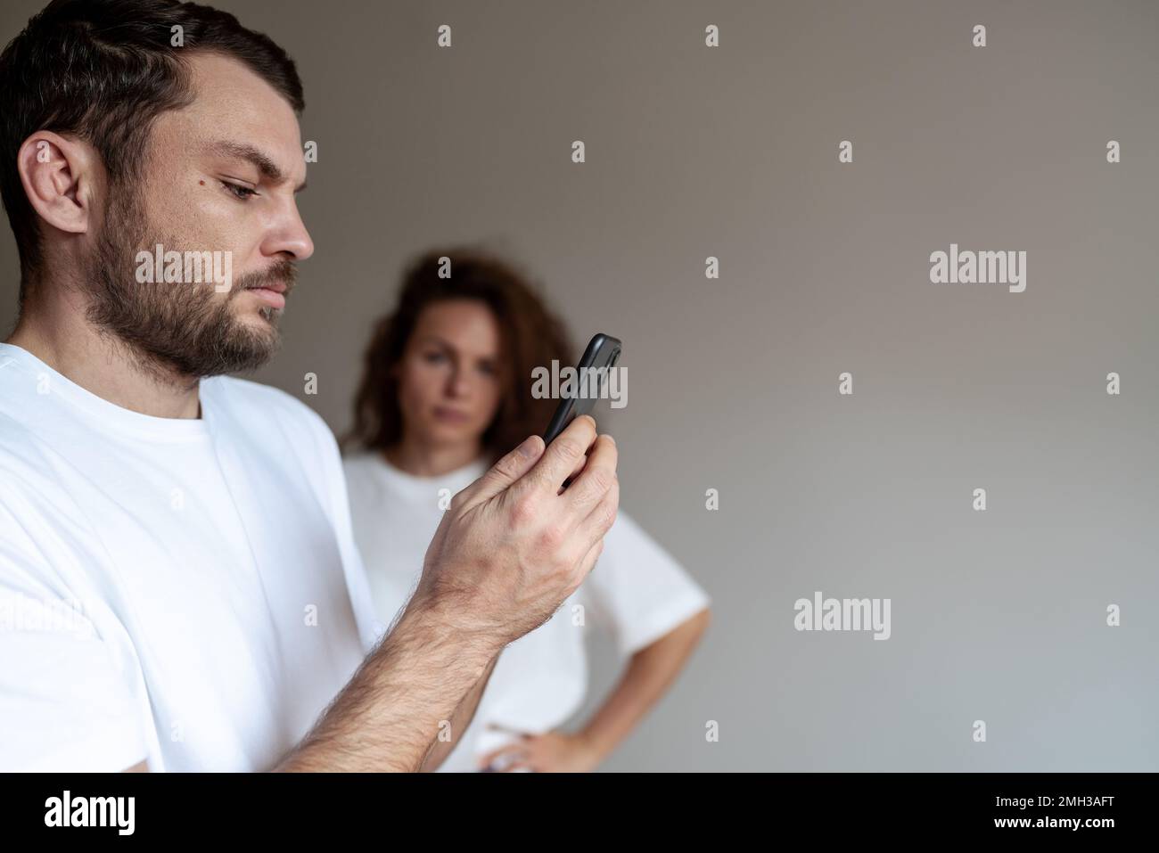 Ein bärtiger Mann liest Nachrichten auf seinem Handy Frau im Hintergrund beobachtet ihren Mann. Stockfoto