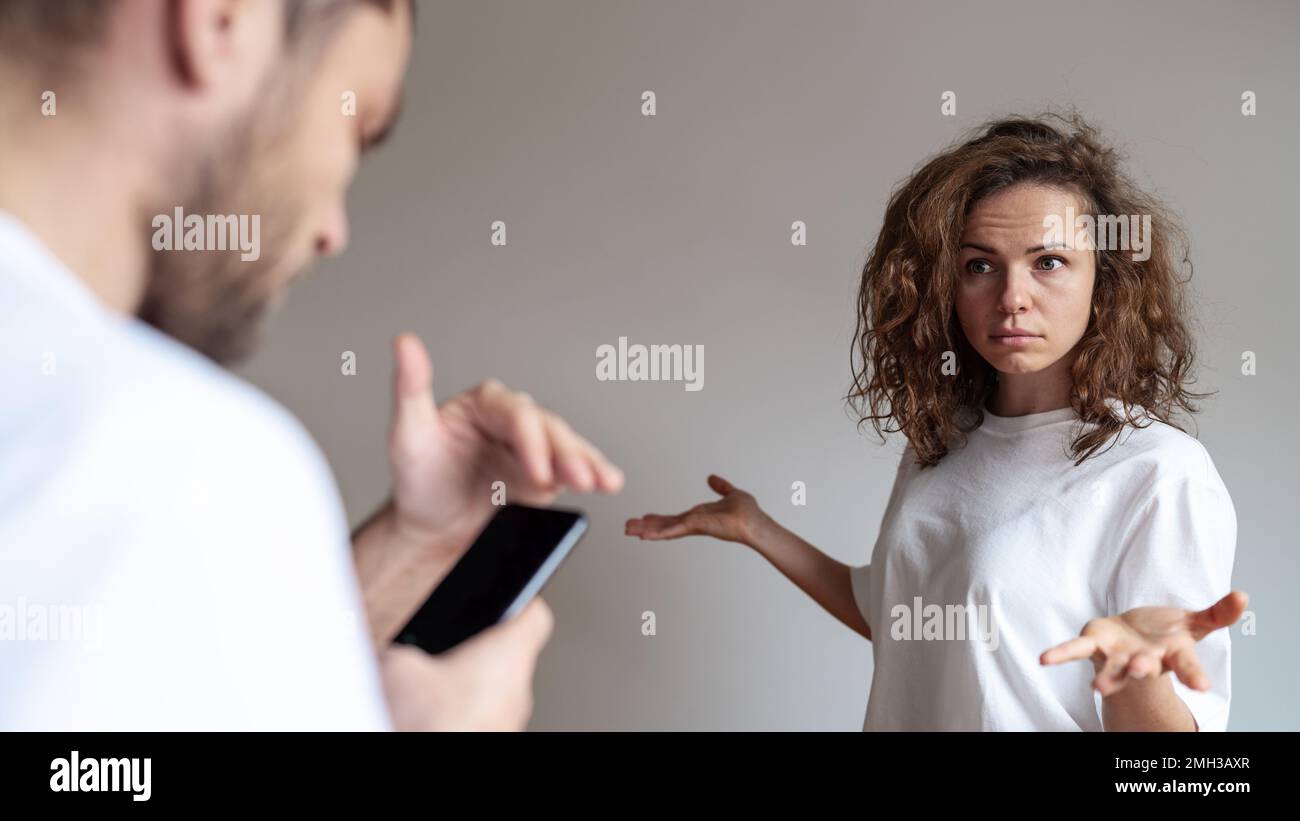Eine verärgerte, lockige Frau sieht einen Ehemann an, der sein Handy benutzt und den Bildschirm mit der Hand bedeckt, Probleme mit der Familie. Untreue, verdächtig Stockfoto