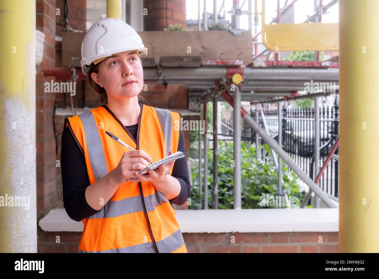 Eine besorgte, konzentrierte Bauingenieurin, die eine Baustelle mit Gerüsten überwacht und dabei einen Schutzhelm und eine orangefarbene psa trägt Stockfoto