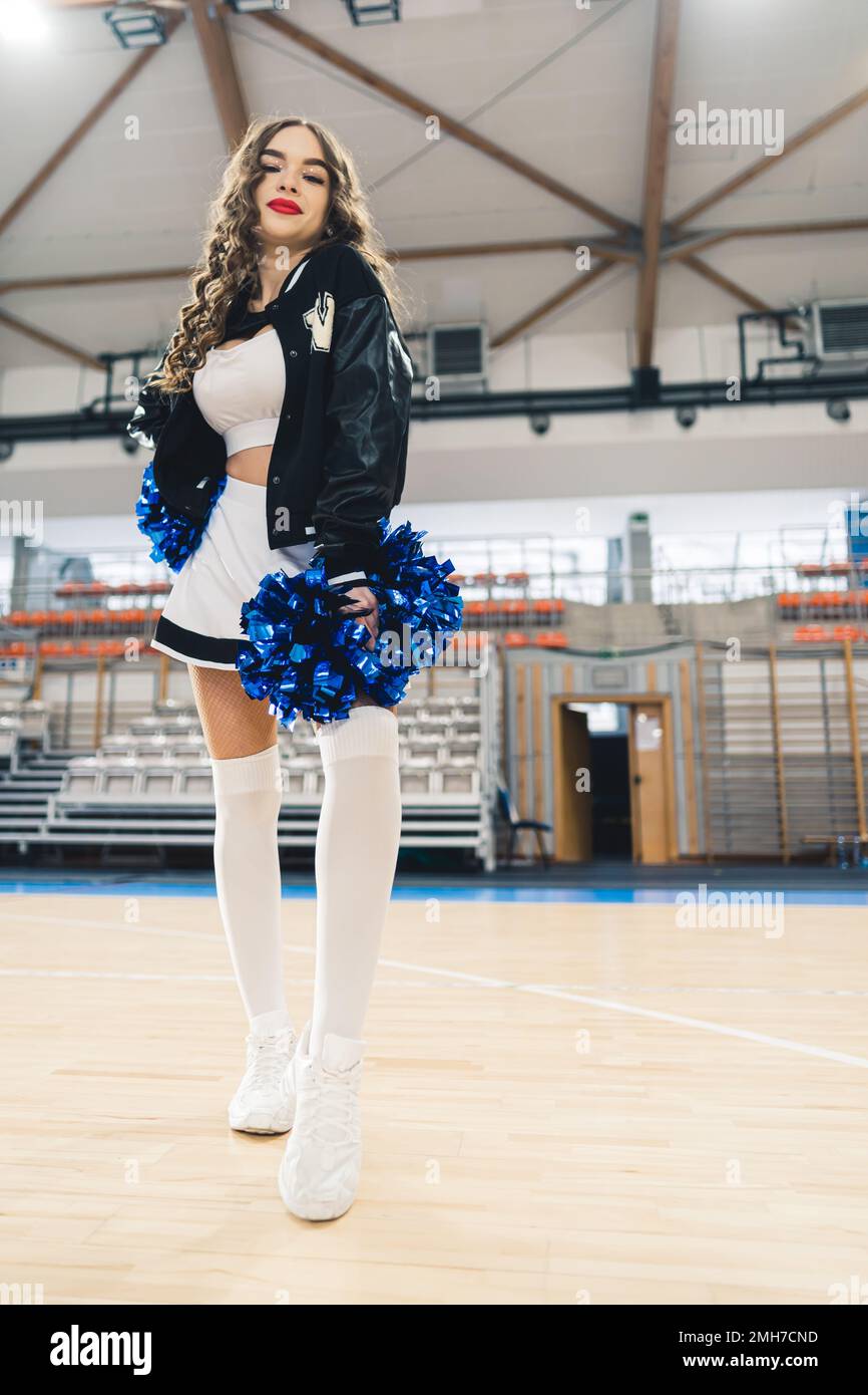 Volle Aufnahme der Cheerleaderin in schwarz-weißer Uniform mit blau glänzenden Pompons. Basketballfeld verschwommen im Hintergrund. Hochwertiges Foto Stockfoto