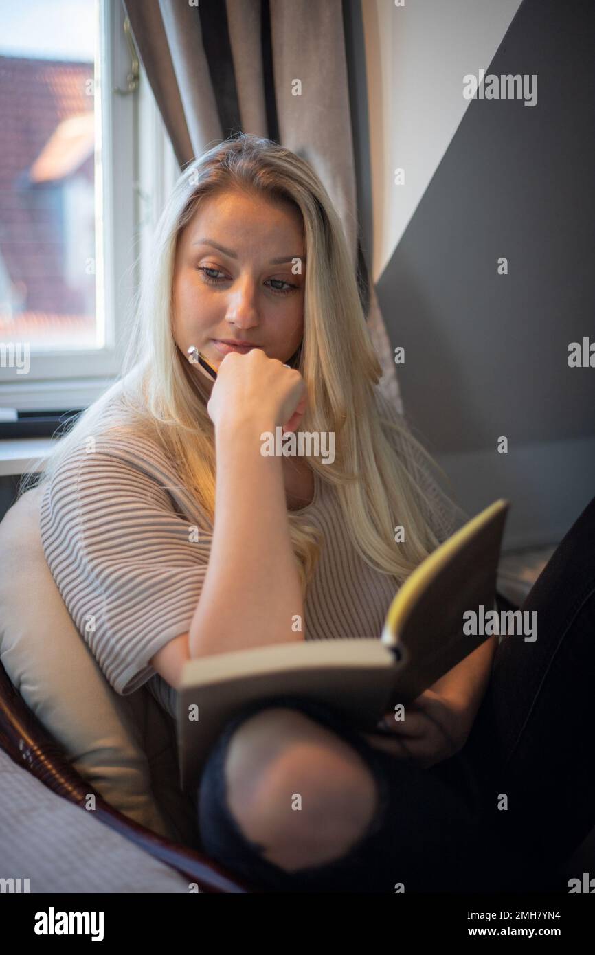 Eine nachdenkliche junge Frau schreibt in ihr Tagebuch, während sie auf dem Sessel sitzt Stockfoto