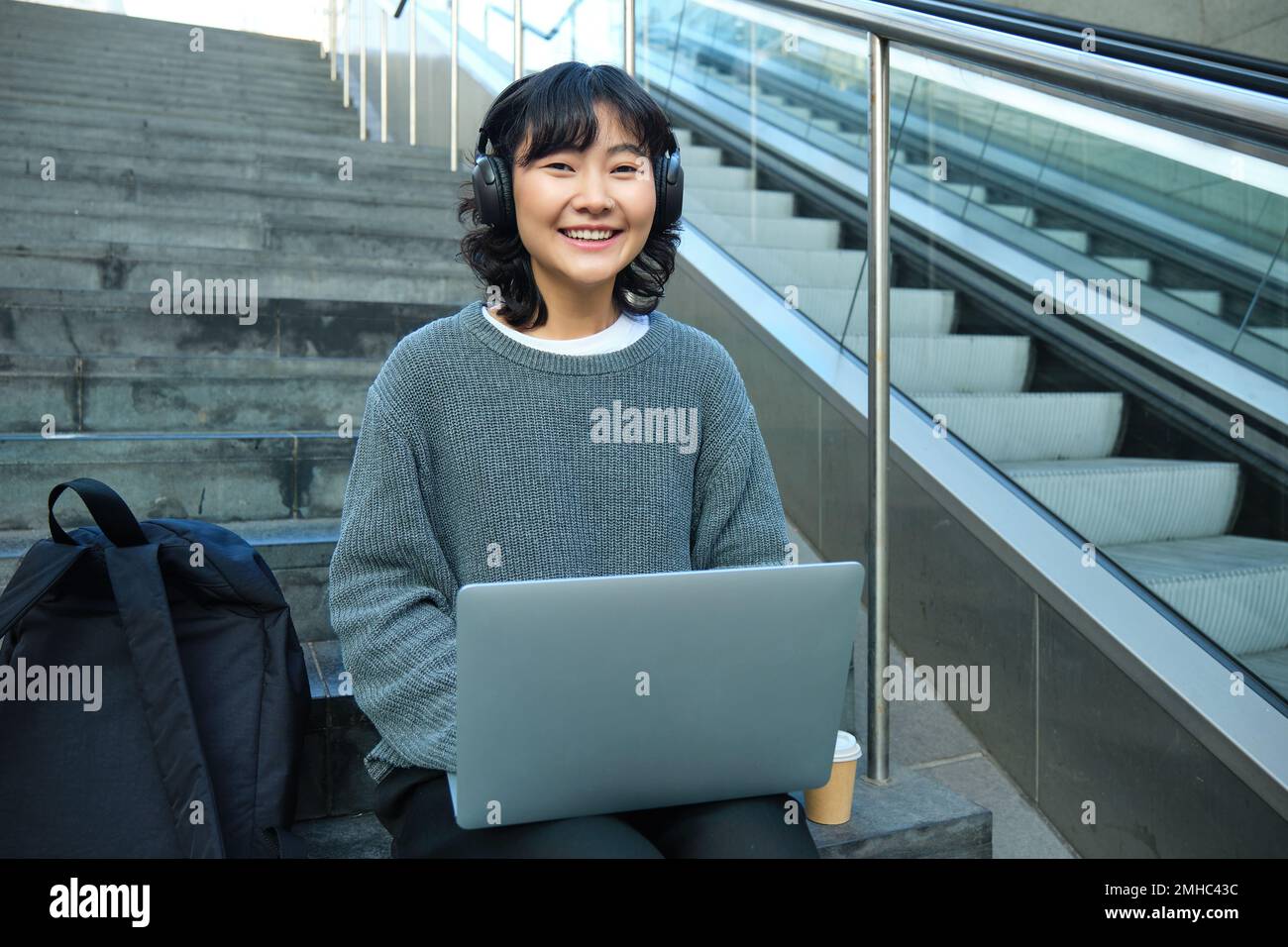 Studentin arbeitet aus der Ferne an einem Projekt, sitzt auf der Treppe, arbeitet auf einem Laptop, hört Musik über Kopfhörer und lächelt Stockfoto