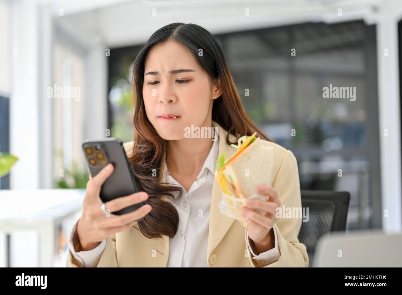 Geschäftige und hungrige asiatische Geschäftsfrau, die ein Sandwich isst, ihr schnelles Frühstück im Büro serviert und gleichzeitig auf die E-Mails von Geschäftskunden antwortet Stockfoto