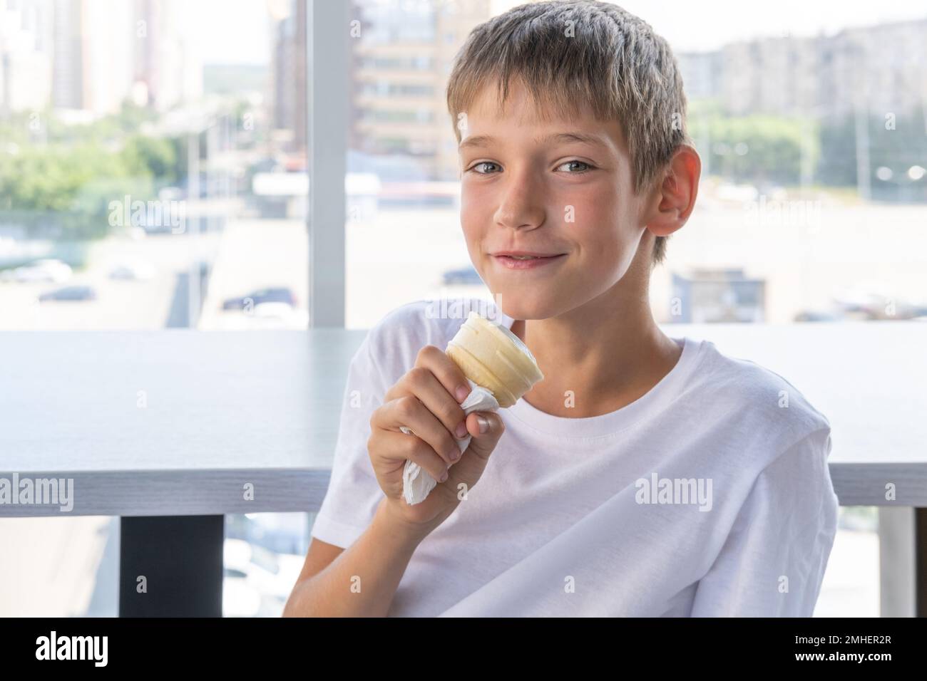 Ein süßer Teenager isst weißes Eis in einem Waffelbecher, sitzt an einem Tisch am Fenster in einem Café. Unscharfer Hintergrund. Sommertag. Ein Kind genießt die Ice Stockfoto