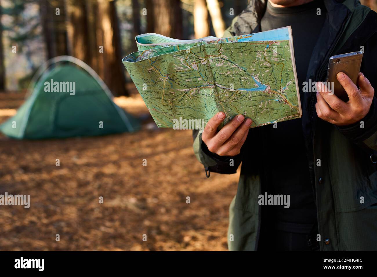 Erwachsener Wanderer mit Rucksack, der Handy und Karte benutzt, im Wald in der Nähe seines Zelts. Wandern älterer Mann in der herbstlichen Natur mit Karte im Freien neben einem Stockfoto