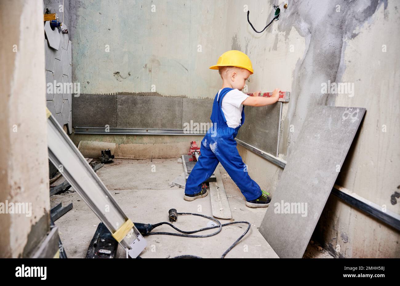 Junge Bauarbeiterin, die die Wandoberfläche mit Wasserwaage in der Wohnung überprüft. Kind trägt Overalls mit waagerechtem Instrument, während es die Wand für Reparaturarbeiten zuhause während der Renovierung vorbereitet. Stockfoto
