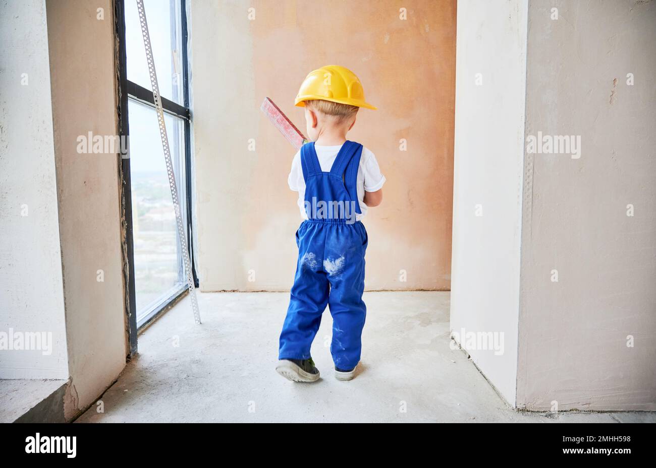 Wandfläche für Kinderkontrolle mit Wasserwaage in der Wohnung. Rückansicht von Kindern im Arbeitsanzug und Sicherheitshelm mit Nivelliergerät bei der Hausrenovierung. Stockfoto
