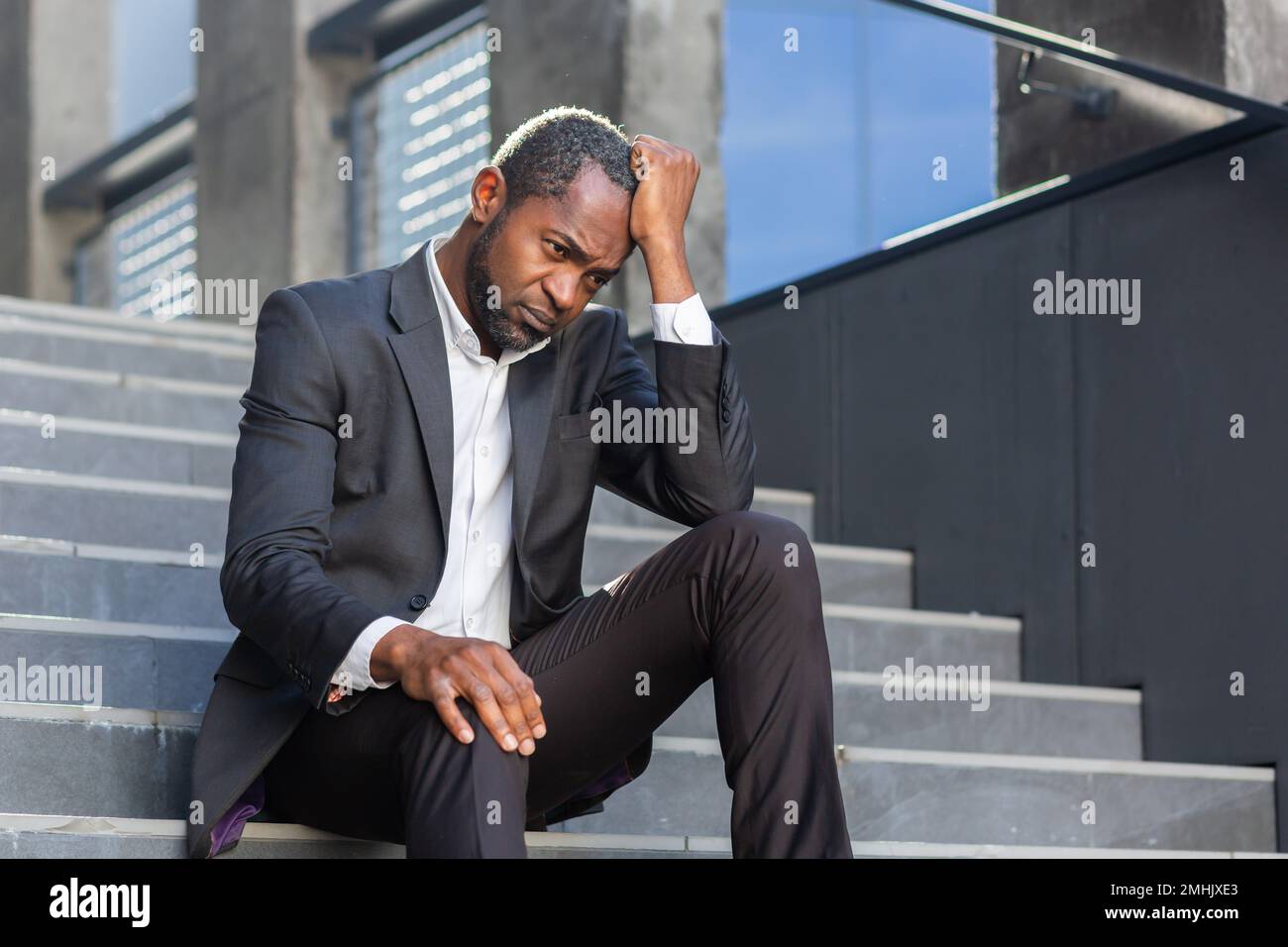 Ein aufgebrachter afroamerikanischer Mann, der vor dem Bürogebäude auf der Treppe sitzt, ein Geschäftsmann, der in Verzweiflung bankrott ist, ein reifer Boss, der in einem Geschäftsanzug im Freien deprimiert ist. Stockfoto