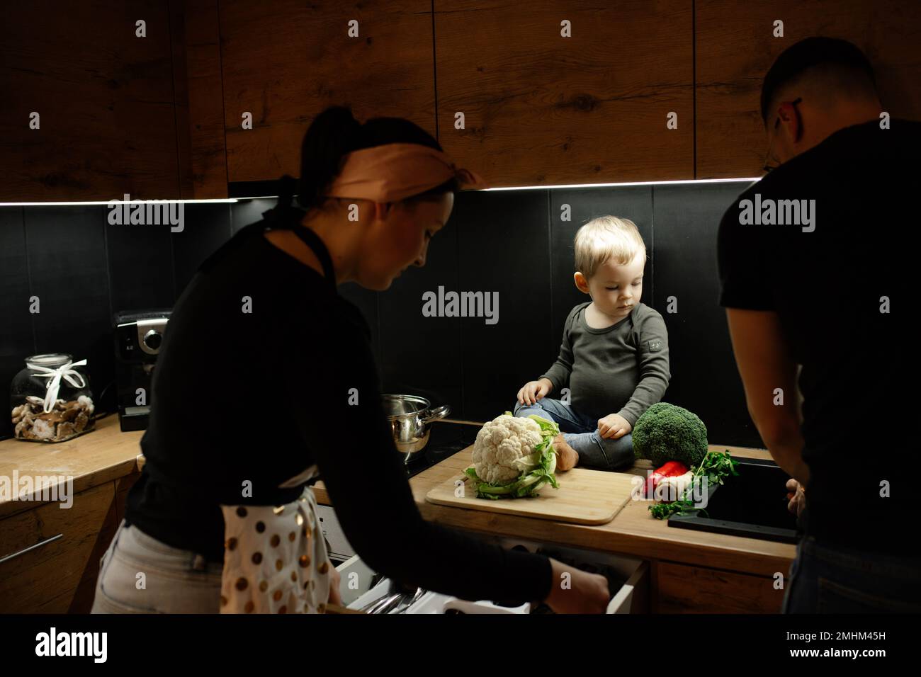 Eltern mit kleinen Kindern kochen in der Küche mit dunklem Licht zu Abend. Bereiten Sie frischen Blumenkohlkopf für das Schneiden an Bord vor. Stockfoto