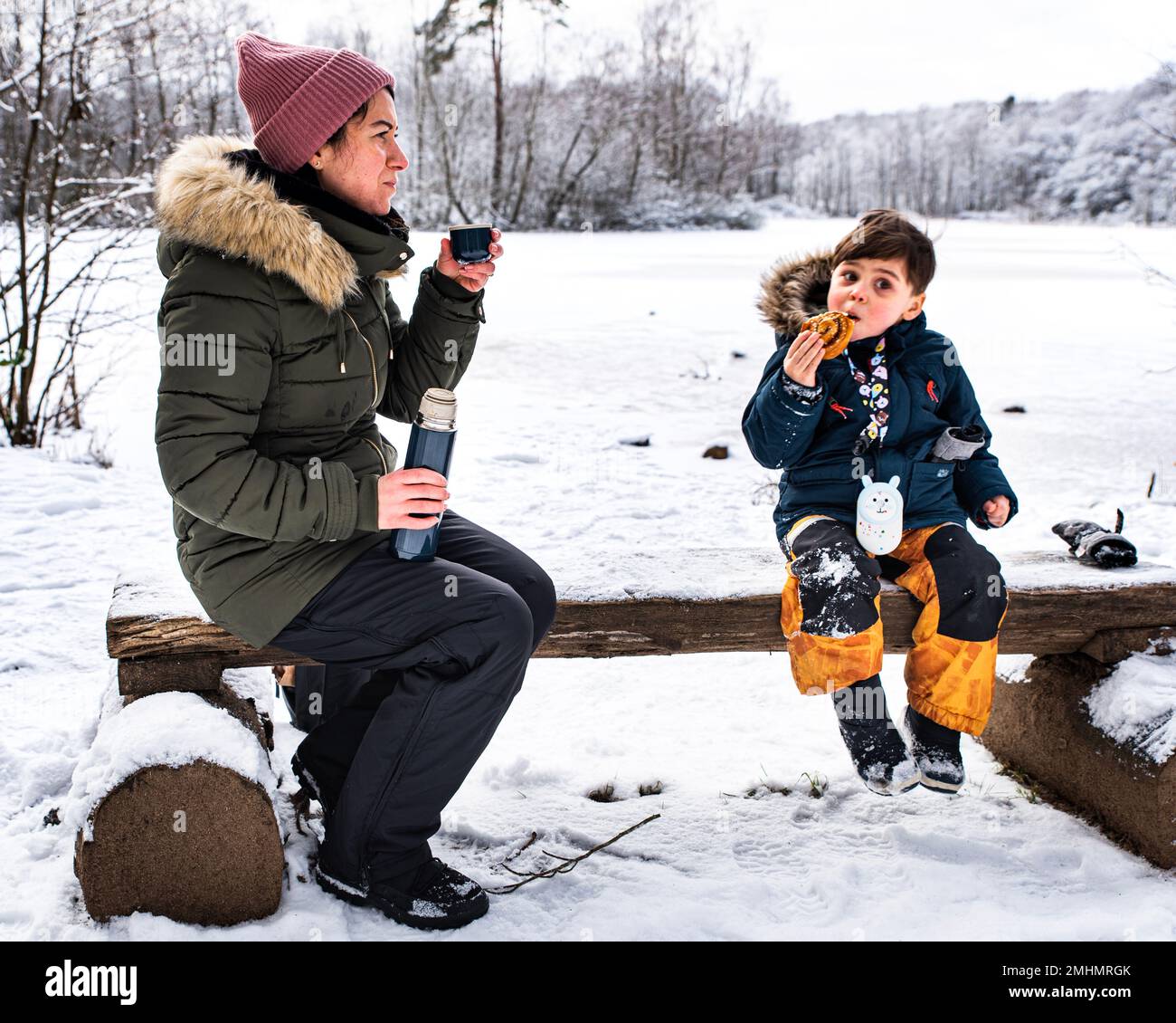 Mutter und Sohn sitzen auf einer Holzbank im Schnee an einem gefrorenen See und genießen eine schwedische Fika mit heißem Kaffee und einer Kanelbular- oder Zimtschnecke Stockfoto