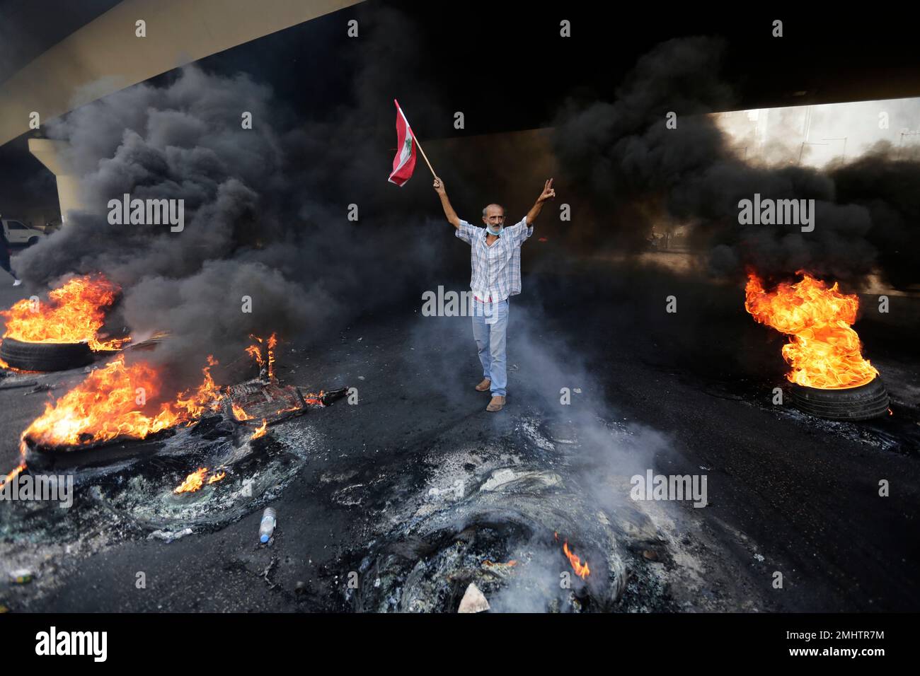An anti-government protester makes victory sign, as he holds a Lebanese national flag and walks fire of tires that sits to block a road during a protest against government's plans to impose new taxes in Beirut, Lebanon, Friday, Oct. 18, 2019. Demonstrators in Lebanon are blocking major roads across the country in a second day of protests against proposed new taxes, which come amid a severe economic crisis. (AP Photo/Hassan Ammar) Stockfoto