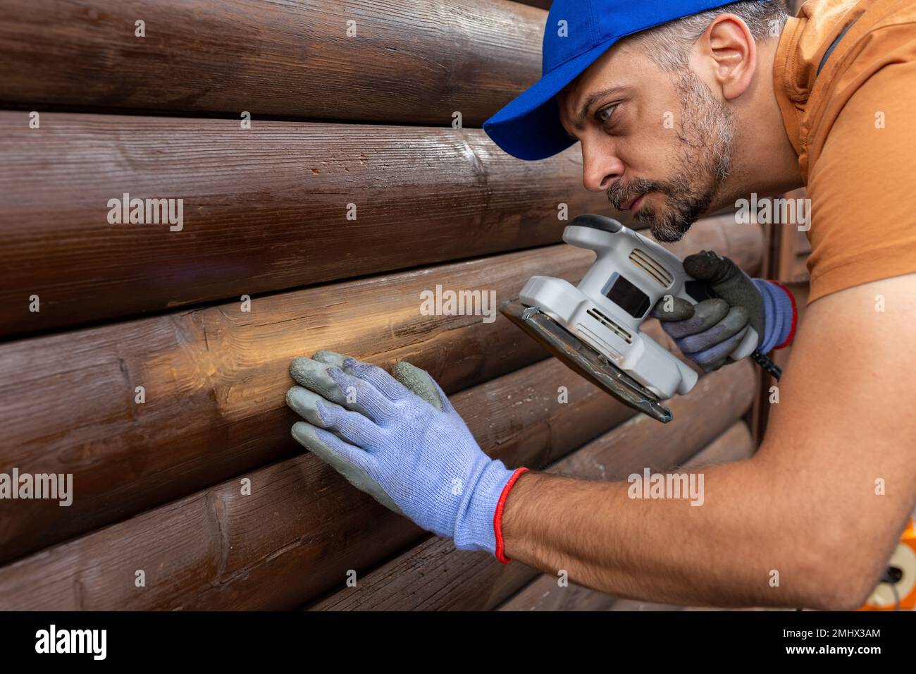 Reparaturarbeiter, der das Holzhaus mit einem Schleifgerät renoviert, bevor er den Fleck zum Wetterschutz aufträgt. Stockfoto