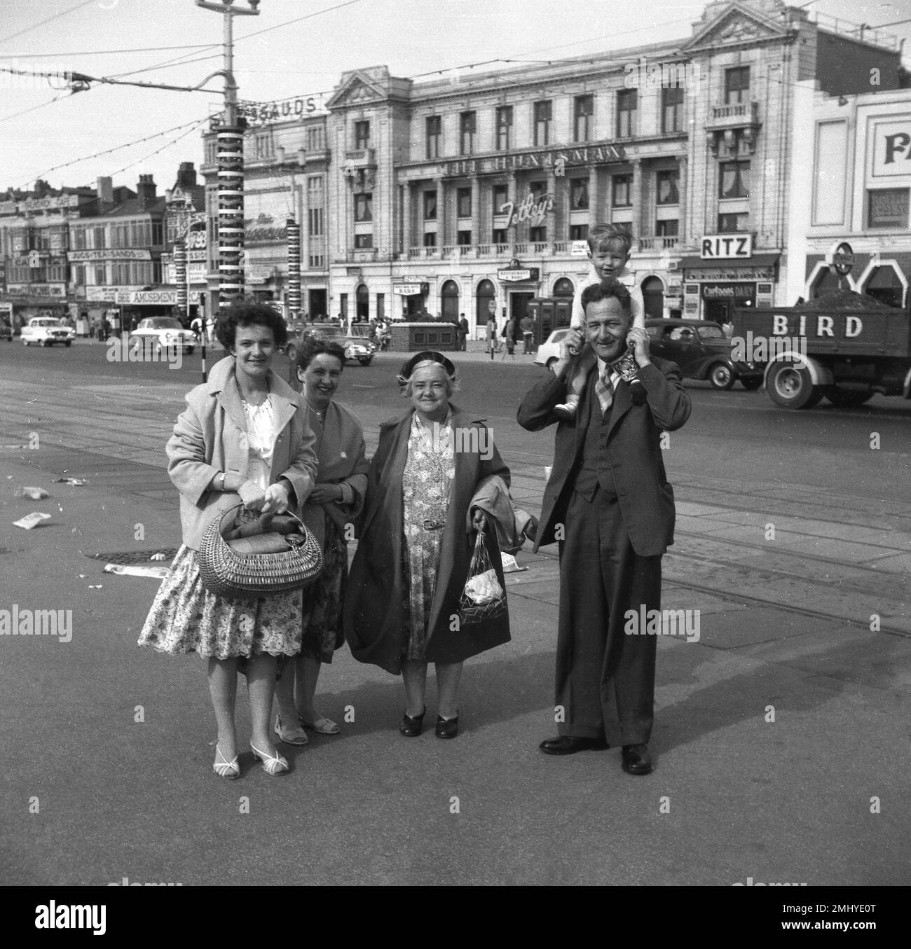 1950er, historisch, eine Arbeiterfamilie, die auf der Promenade in Blackpool, England, für ein Foto steht. Auf der Straße dahinter, auf der Royal Princes Parade, Autos der Epoche. Huntsman, Ritz, Tassauds (Wachsfiguren) sind Namen auf den Gebäuden. Die Familie ist formell gekleidet, da die Leute in dieser Zeit im Urlaub sind, trägt der Gentleman mit einem kleinen Jungen auf seinen Schultern ein dreiteiliges Sit & Krawatte und die Frauen in Blumenkleidern und langen Mänteln. Stockfoto