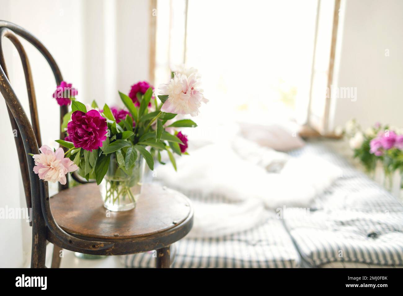 Nahaufnahme von Pfingstrosen in einer Vase auf einem alten Holzstuhl. Rustikale Einrichtung mit Matratzen, Kissen und Blumen in der Nähe eines offenen Fensters. Atmosphärisch m Stockfoto