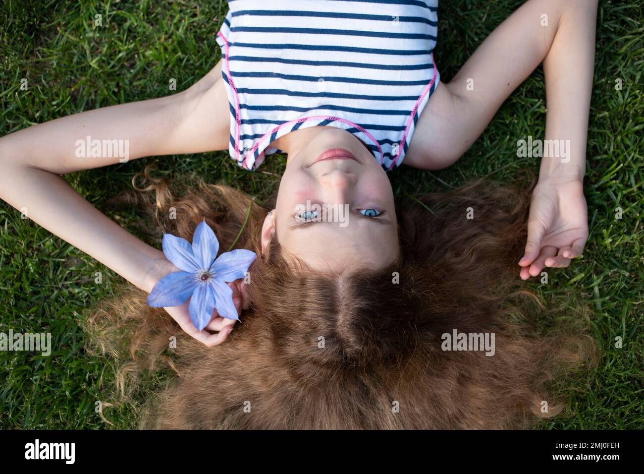 Foto eines süßen Mädchens, 11-12 Jahre alt, traumhaft auf dem Gras liegt, mit ausgestreckten Haaren, sieht nach oben. Blick von oben. Positives Lächeln, Haarpflege, Entspannen Stockfoto