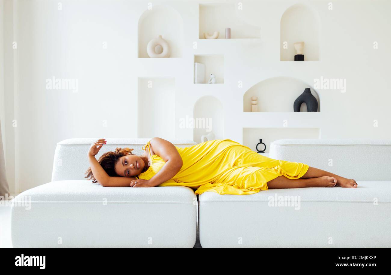 Eine wunderschöne, kurvige, übergroße, junge afto amerikanische Frau in einem gelben Kleid liegt zu Hause auf dem Sofa. Körperpositive, stilvolle, lächelnde Frau hat eine entspannte Zeit. Bsb Stockfoto