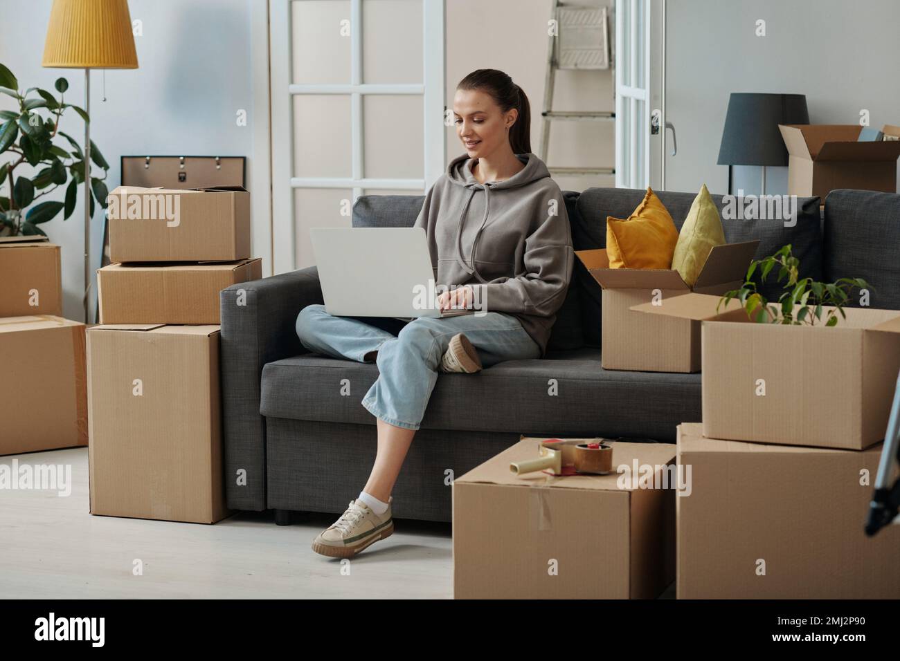 Mädchen sitzt auf dem Sofa und sucht mit dem Laptop nach Spielfiguren, während sie in ein neues Haus zieht Stockfoto