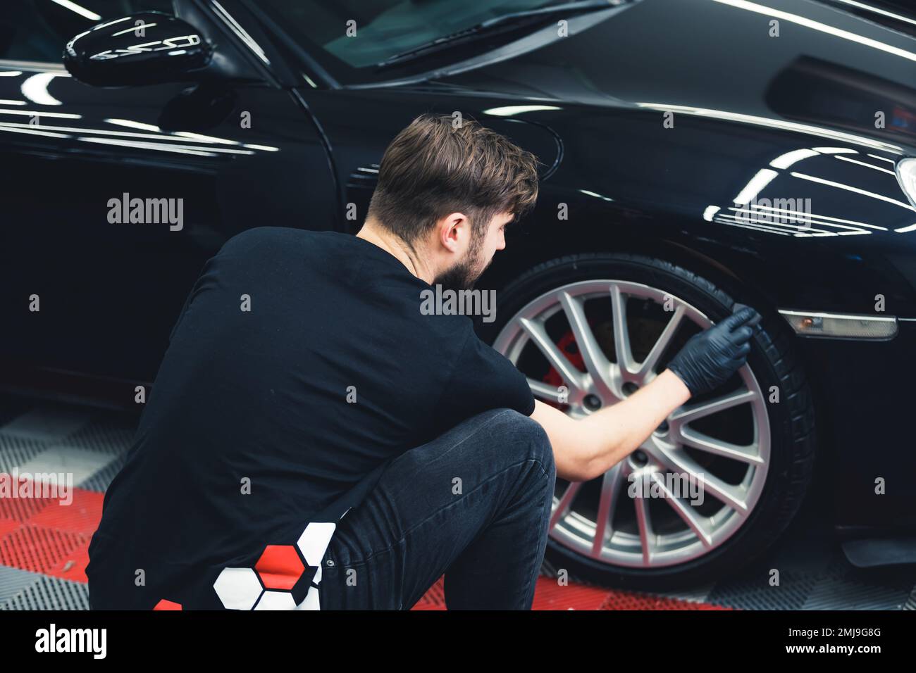 Rückansicht eines weißen Mannes mit schwarzen Handschuhen, mit Schwamm gebeugte, schwarze Autoreifen. Professionelle Autoverbesserung in einer Garage. Horizontaler Innenschuss. Hochwertiges Foto Stockfoto