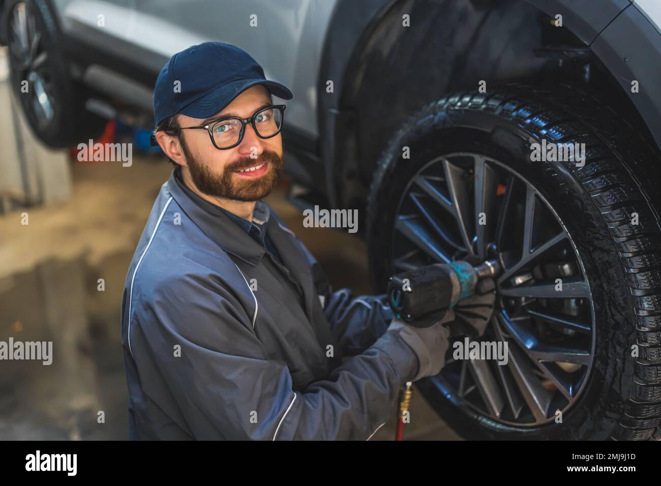 Aufnahme eines weißen Mechanikers, der die Kamera anlächelt, während er ein Autorad mit einem Schlagschrauber verschraubt. Werkstattkonzept. Hochwertiges Foto Stockfoto