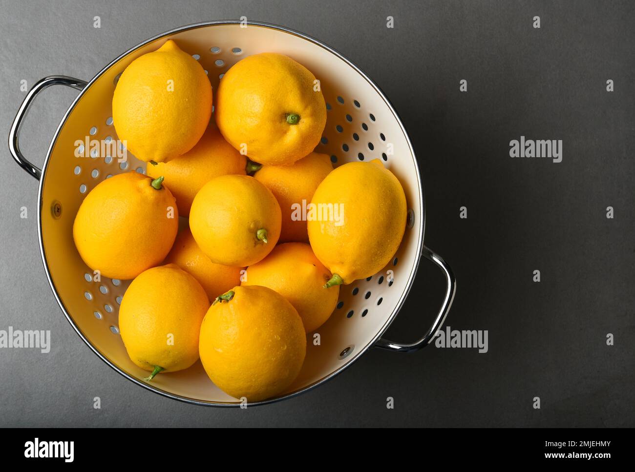 Frisch gepflückte Zitronen in einem weißen Sieb auf einer grauen Schieferfliese mit starkem Seitenlicht. Aufnahme aus hohem Winkel mit Kopierbereich auf der rechten Seite. Stockfoto