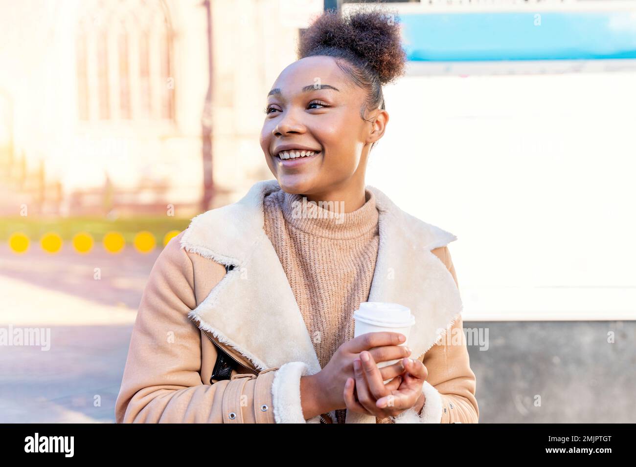 Fröhliche Afrikanerin hält eine Tasse Kaffee. Eine lächelnde braune Dame in braunem Pullover, die auf eine Straßenbahn wartet. Stockfoto