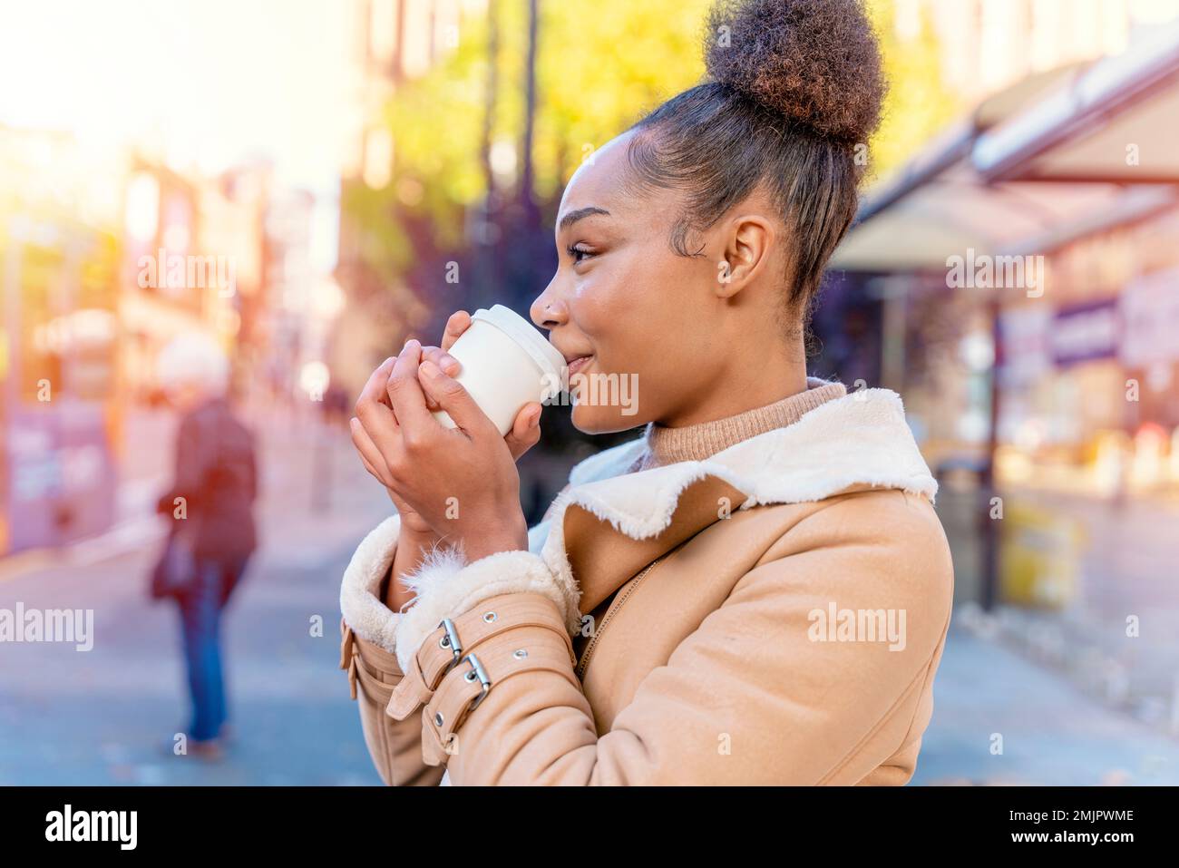 Fröhliche Afrikanerin hält eine Tasse Kaffee. Eine lächelnde braune Dame in braunem Pullover, die auf eine Straßenbahn wartet. Stockfoto