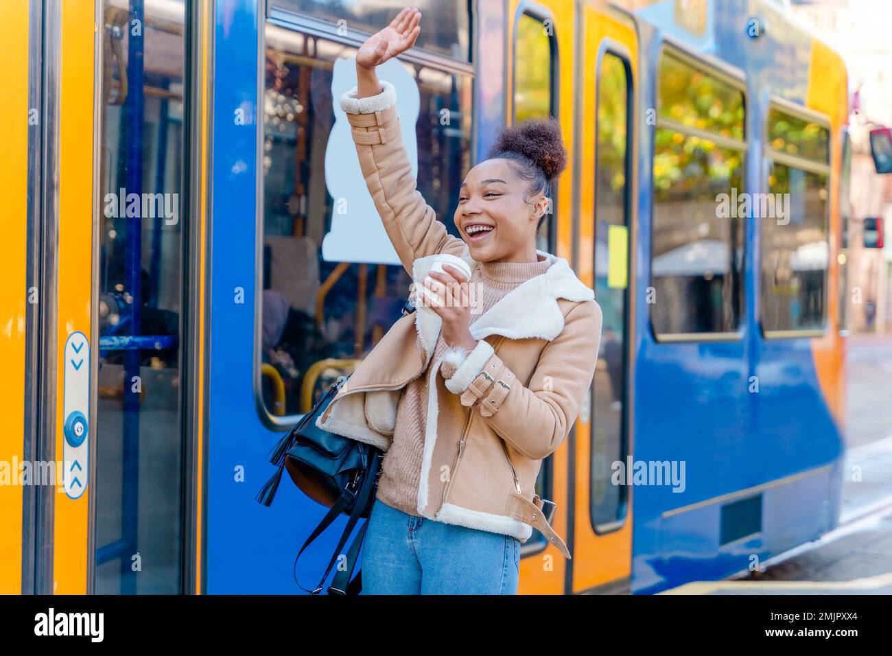 Fröhliche Afrikanerin hält eine Tasse Kaffee. Eine lächelnde braune Dame in braunem Pullover, die auf eine Straßenbahn wartet. Stockfoto