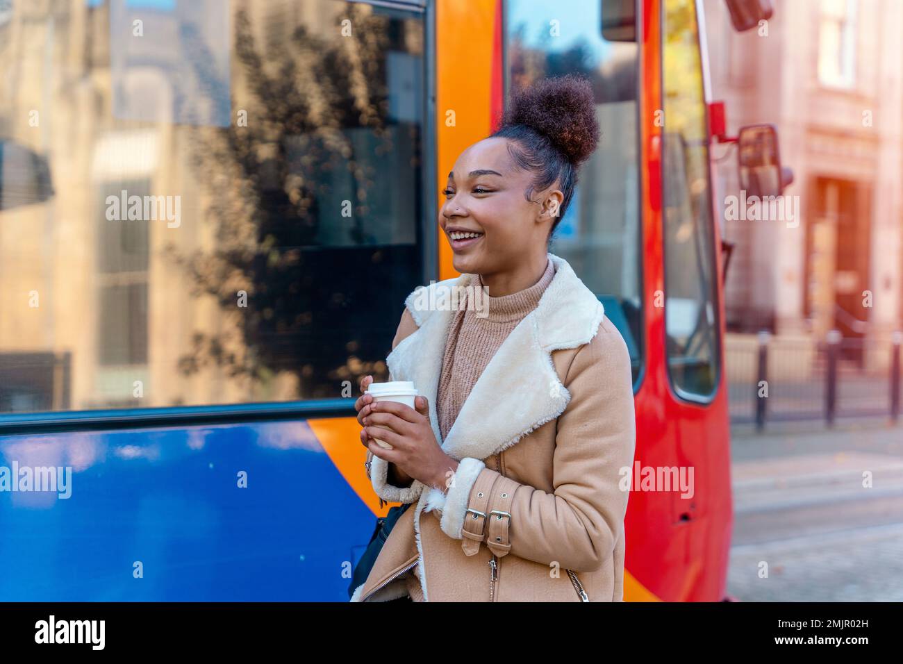 Fröhliche Afrikanerin hält eine Tasse Kaffee. Eine lächelnde braune Dame in braunem Pullover, die auf eine Straßenbahn wartet. Stockfoto