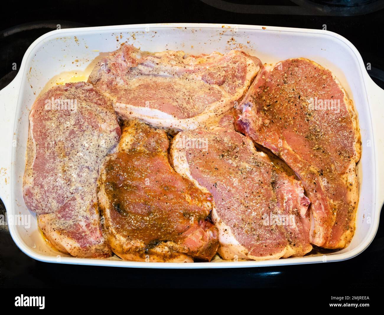 Mehrere rohe Schweinekoteletts mit Gewürzen und Öl in Keramik-Backform auf dem Herd der Küche Stockfoto