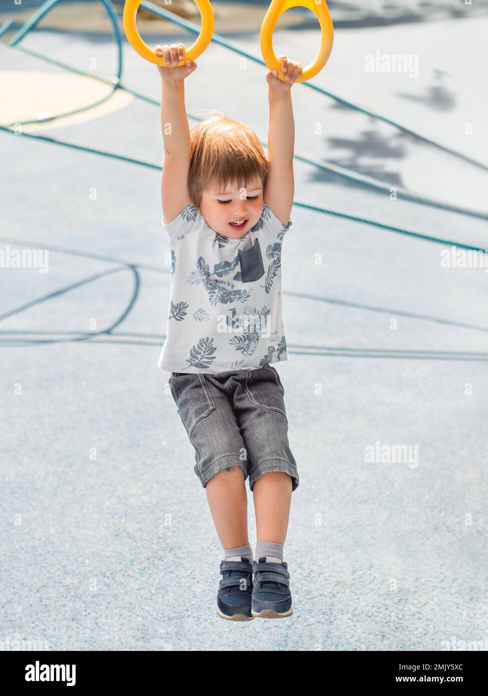 Der kleine Junge trainiert auf dem öffentlichen Sportgelände mit blauem Pflaster. Der Junge macht Übungen an Sportringen. Aktive Freizeitaktivitäten im Freien. Stockfoto