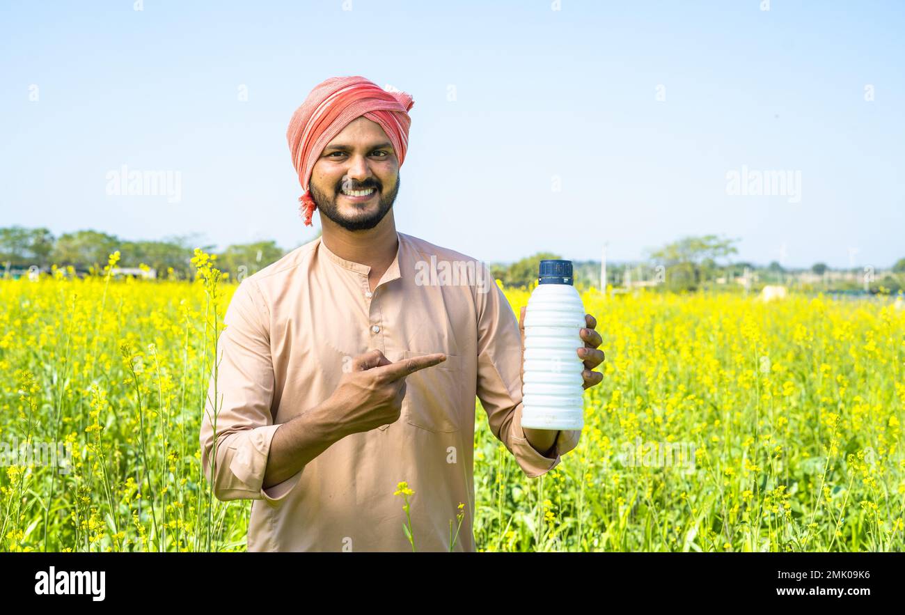Selbstbewusster Junglandwirt, der die Düngemittelflasche mit dem Finger zeigt, während er auf das Ackerland in die Kamera schaut - Konzept der Produktempfehlung Stockfoto
