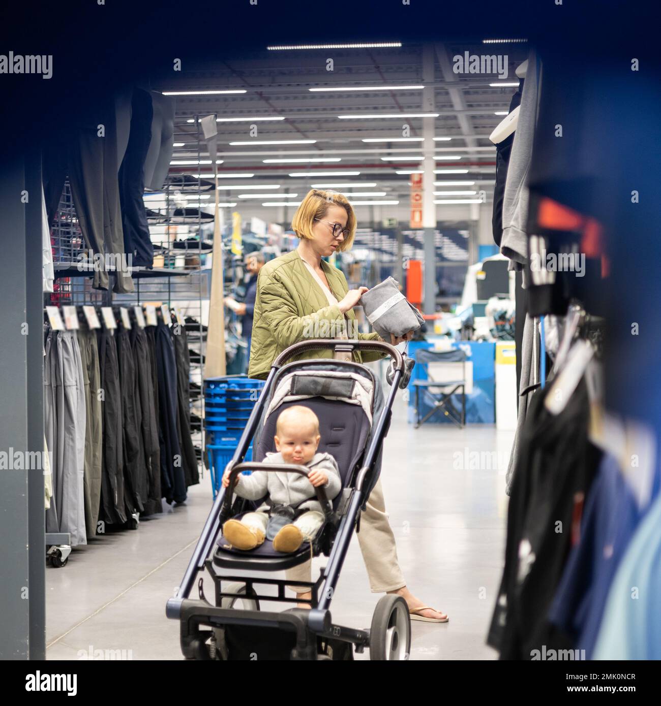 Lässig gekleidete Mutter, die in der Sportabteilung des Supermarkts Sportschuhe und Bekleidungsartikel aussucht, mit ihrem kleinen Jungen im Kinderwagen Stockfoto
