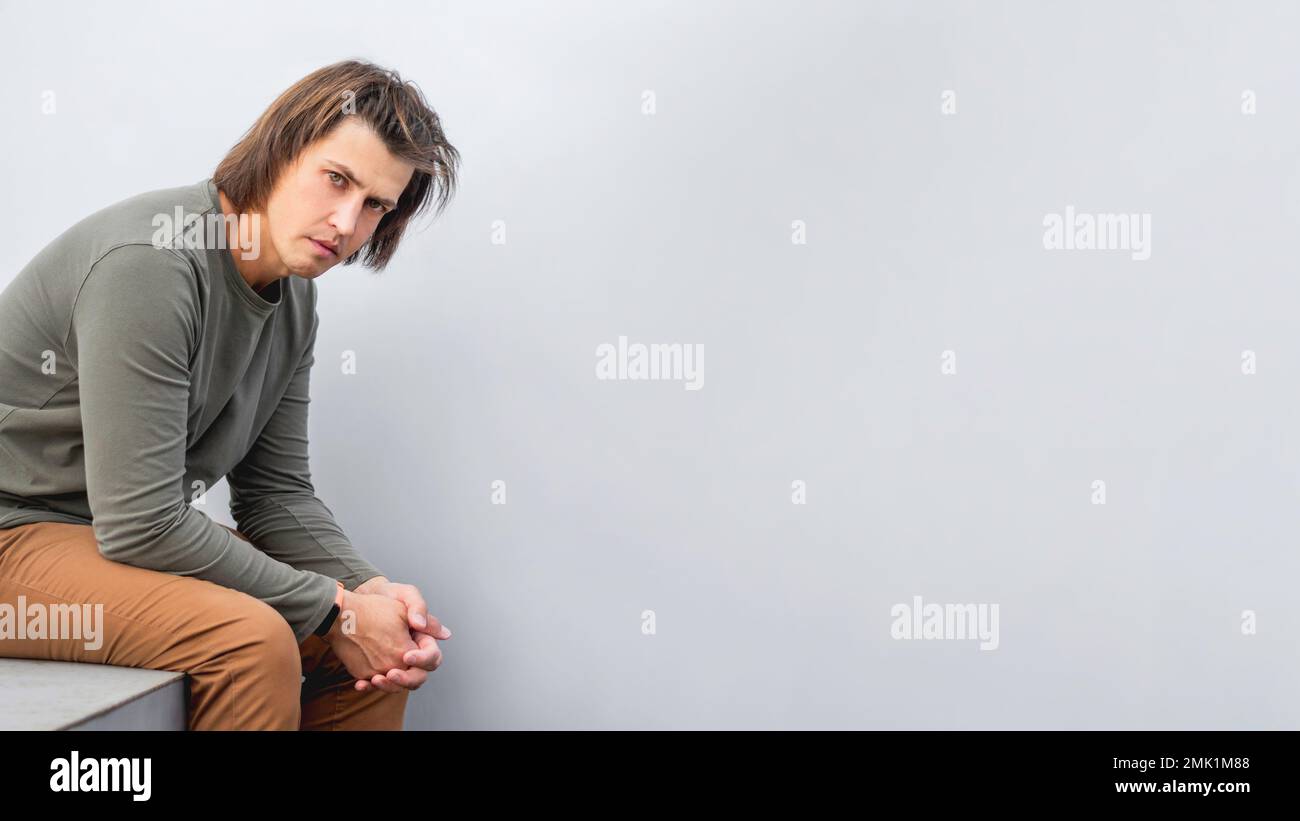 Ein Mann der Generation Y mit langen Haaren und angespanntem Look sitzt an der grauen Wand. Ein seriöser Mann in erdgetönter Freizeitkleidung starrt vor die Kamera. Speicherplatz kopieren. Stockfoto