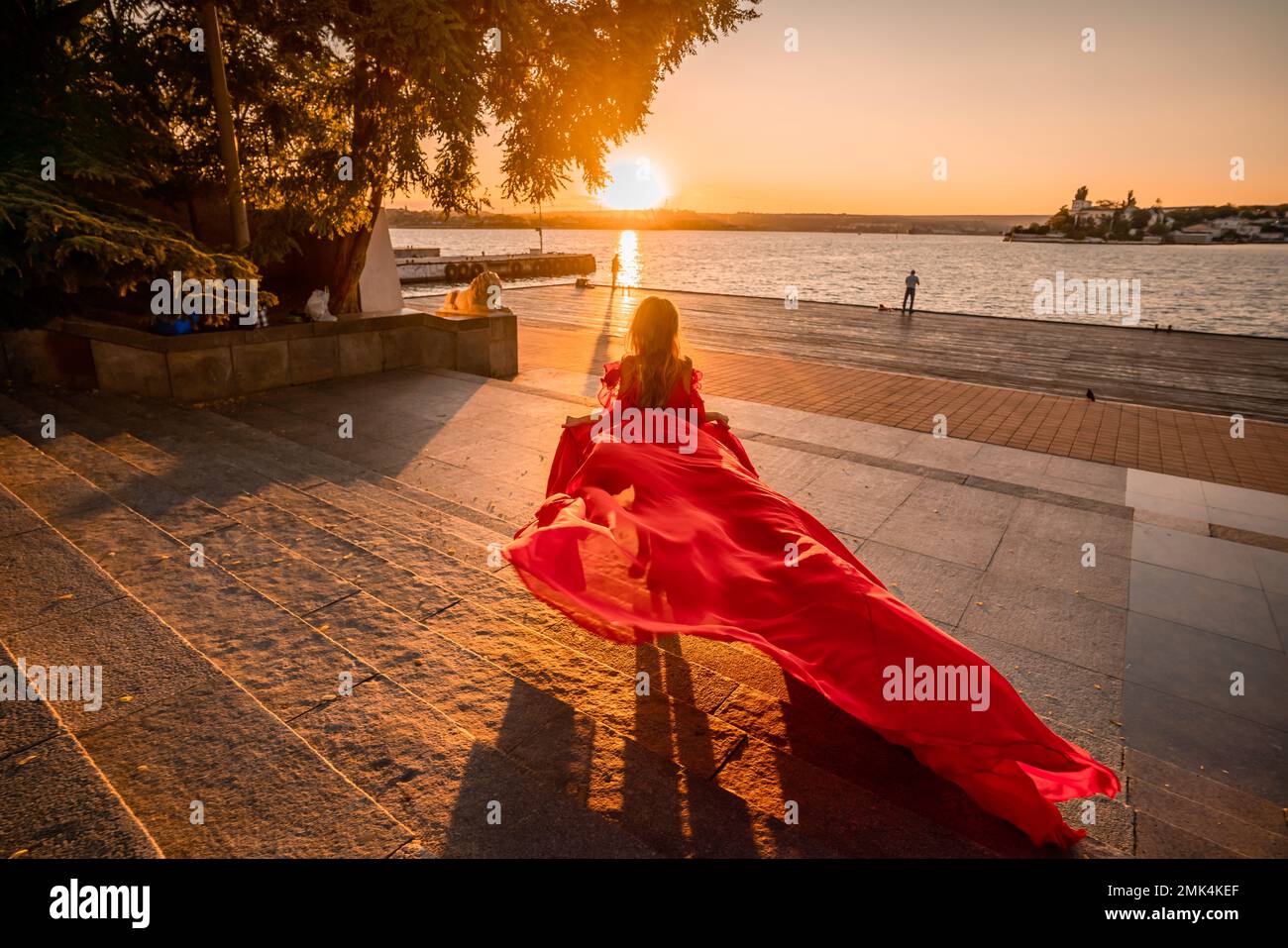 Rotes Kleid bei Sonnenaufgang. Eine Frau in einem langen roten Kleid vor dem Hintergrund des Sonnenaufgangs, helles goldenes Licht der Sonnenstrahlen. Der Begriff der Weiblichkeit Stockfoto