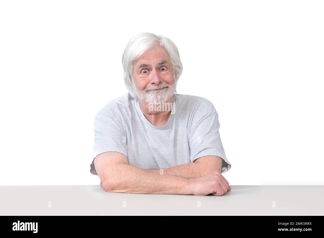 Horizontale Aufnahme eines albernen alten Mannes, der mit gekreuzten Armen an einem Tisch sitzt. Isoliert auf weiß. Viel Platz zum Kopieren. Stockfoto