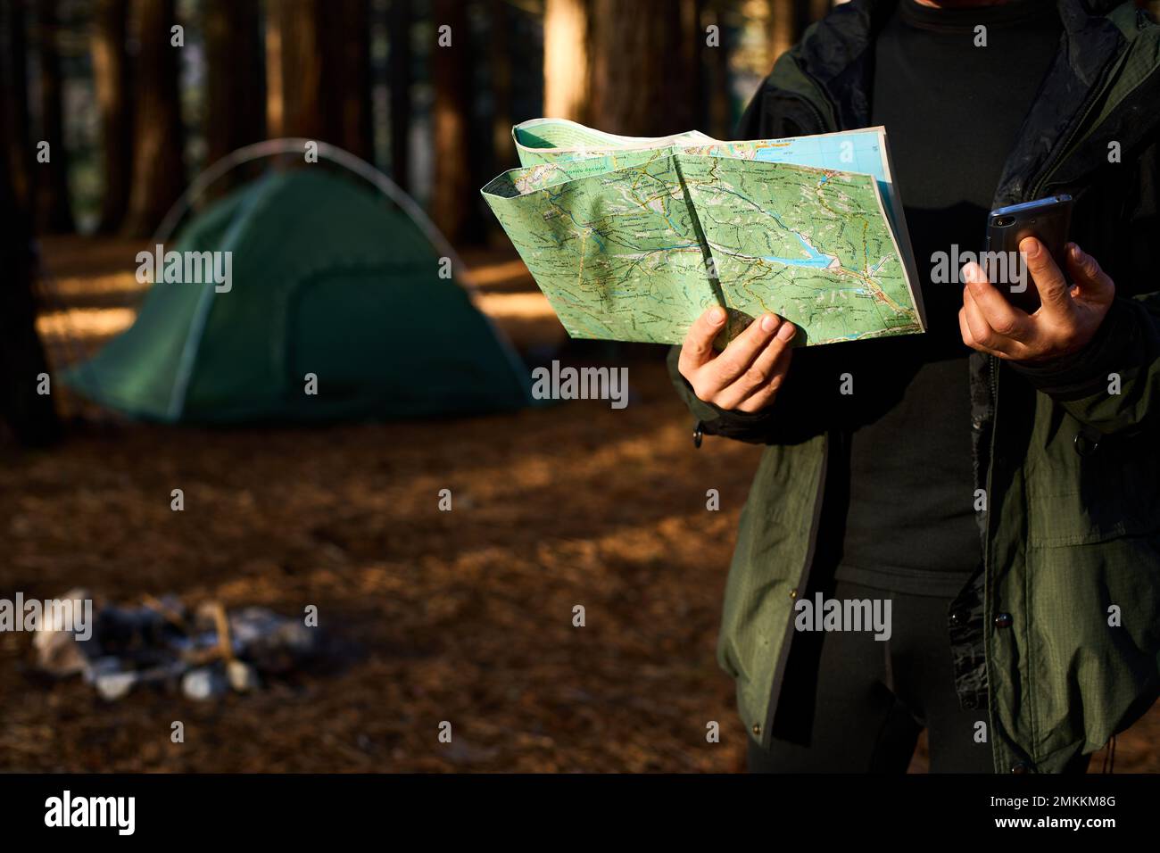 Erwachsener Wanderer mit Rucksack, der Handy und Karte benutzt, im Wald in der Nähe seines Zelts. Wandern älterer Mann in der herbstlichen Natur mit Karte im Freien neben einem Stockfoto