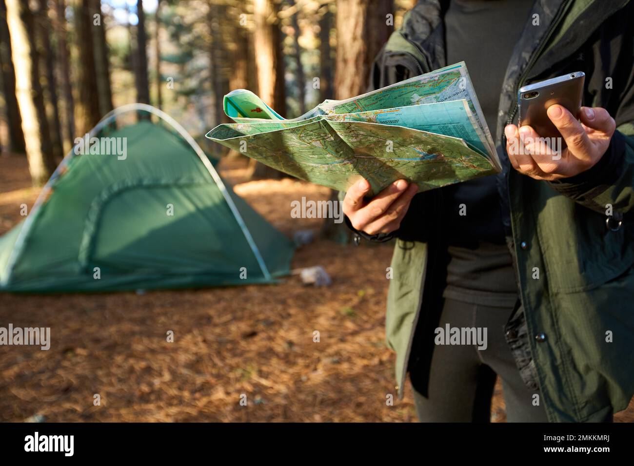 Erwachsener Wanderer mit Rucksack, der Handy und Karte benutzt, im Wald in der Nähe seines Zelts. Wandern älterer Mann in der herbstlichen Natur mit Karte im Freien neben einem Stockfoto