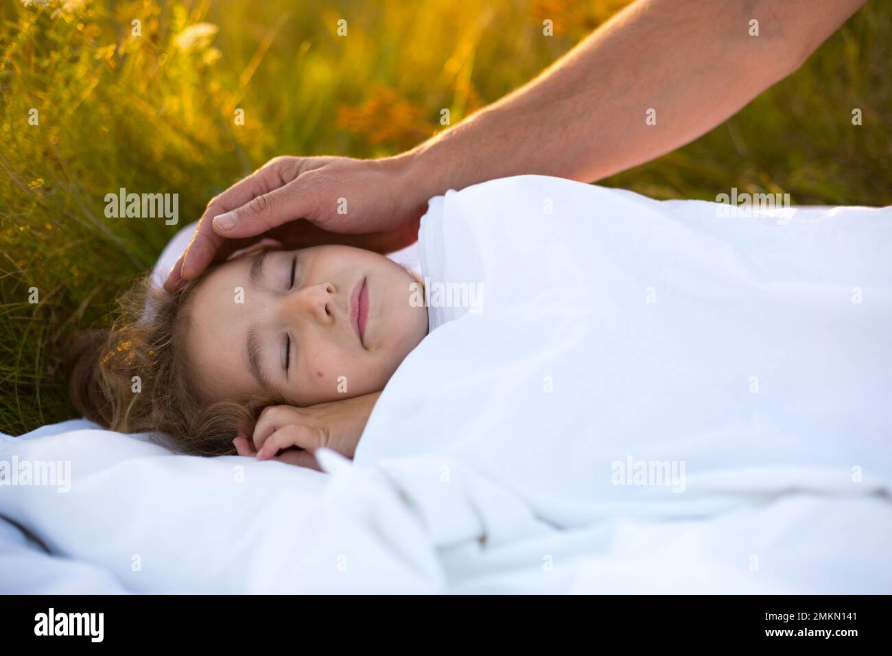 Mädchen schläft auf weißem Bett im Gras, frische Luft. Dads Hand klopft ihm sanft auf den Kopf. Pflege, Schutz, Internationaler Kindertag, Mückenstiche Stockfoto
