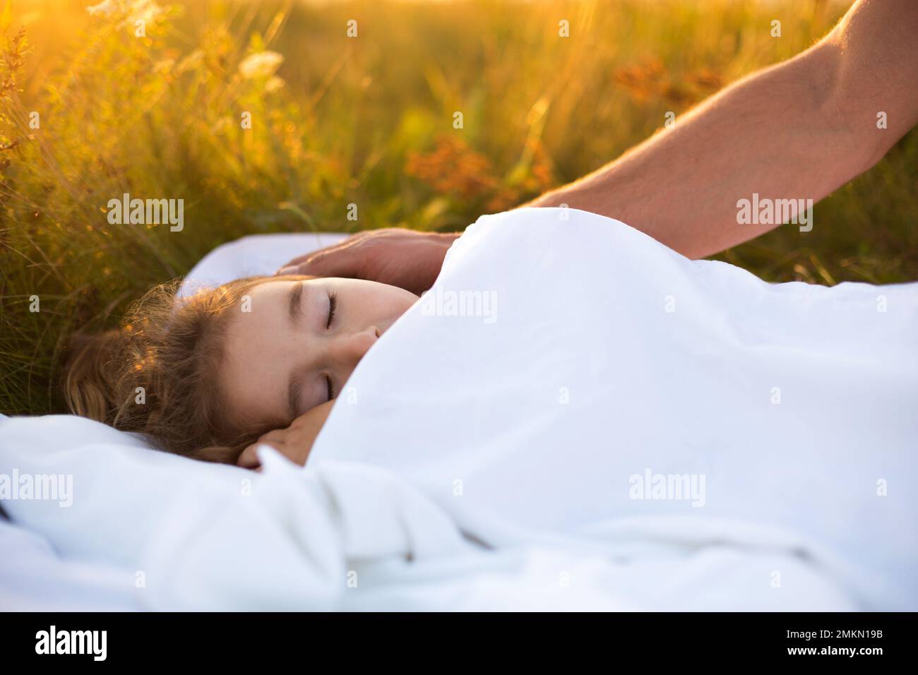 Mädchen schläft auf weißem Bett im Gras, frische Luft. Dads Hand klopft ihm sanft auf den Kopf. Pflege, Schutz, Internationaler Kindertag, Mückenstiche Stockfoto