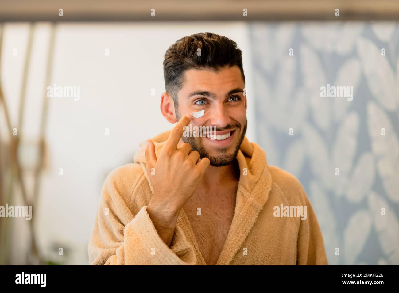 Glücklicher Mann im Bademantel, der lächelt und Creme auf die Wange aufträgt, während er bei der Hautpflege zu Hause in den Spiegel schaut Stockfoto