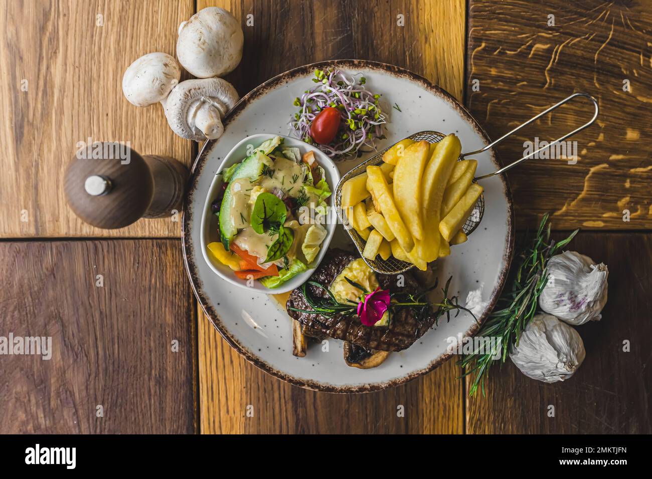 Rinderstriploin-Steak mit Pommes Frites in einem Metallkorb und Salat. Rustikaler Holztisch, mit Muschrroms und Knoblauch dekoriert. Horizontales Innenbild von oben nach unten. Hochwertiges Foto Stockfoto