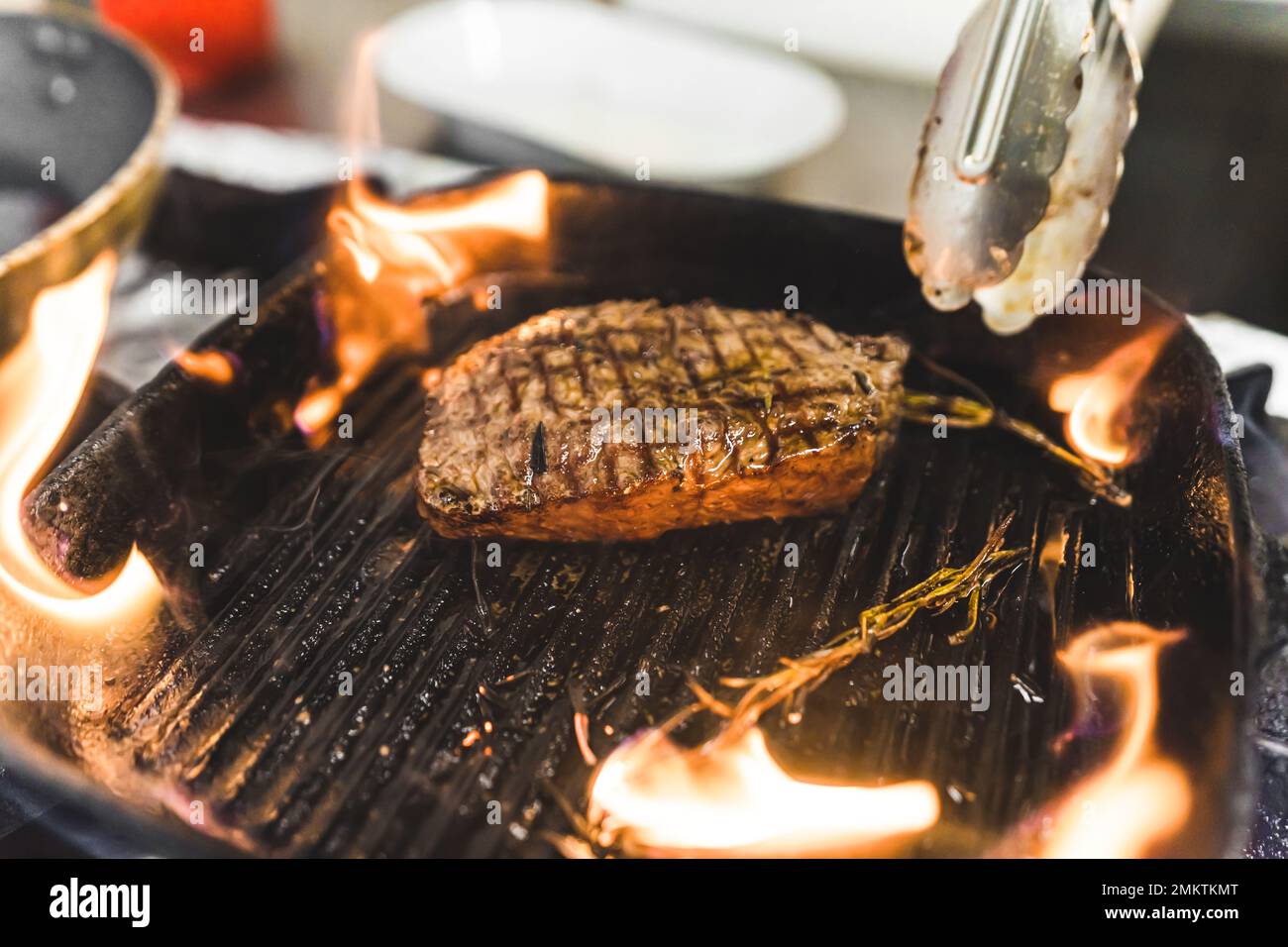 Rinderstriploin-Steak auf einer Grillpfanne mit Kräutern zubereitet. Brennende Flammen. Speisezubereitung in der Küche des Restaurants. Horizontale Indoor-Aufnahme. Hochwertiges Foto Stockfoto