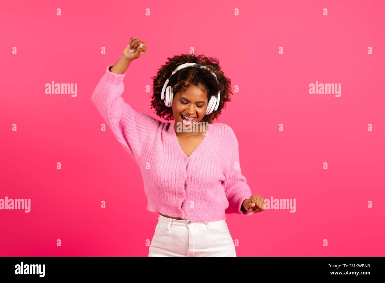 Lächelnde junge afroamerikanische lockige Dame in lässigen und kabellosen Kopfhörern mit einer Zahnspange zum Tanzen Stockfoto