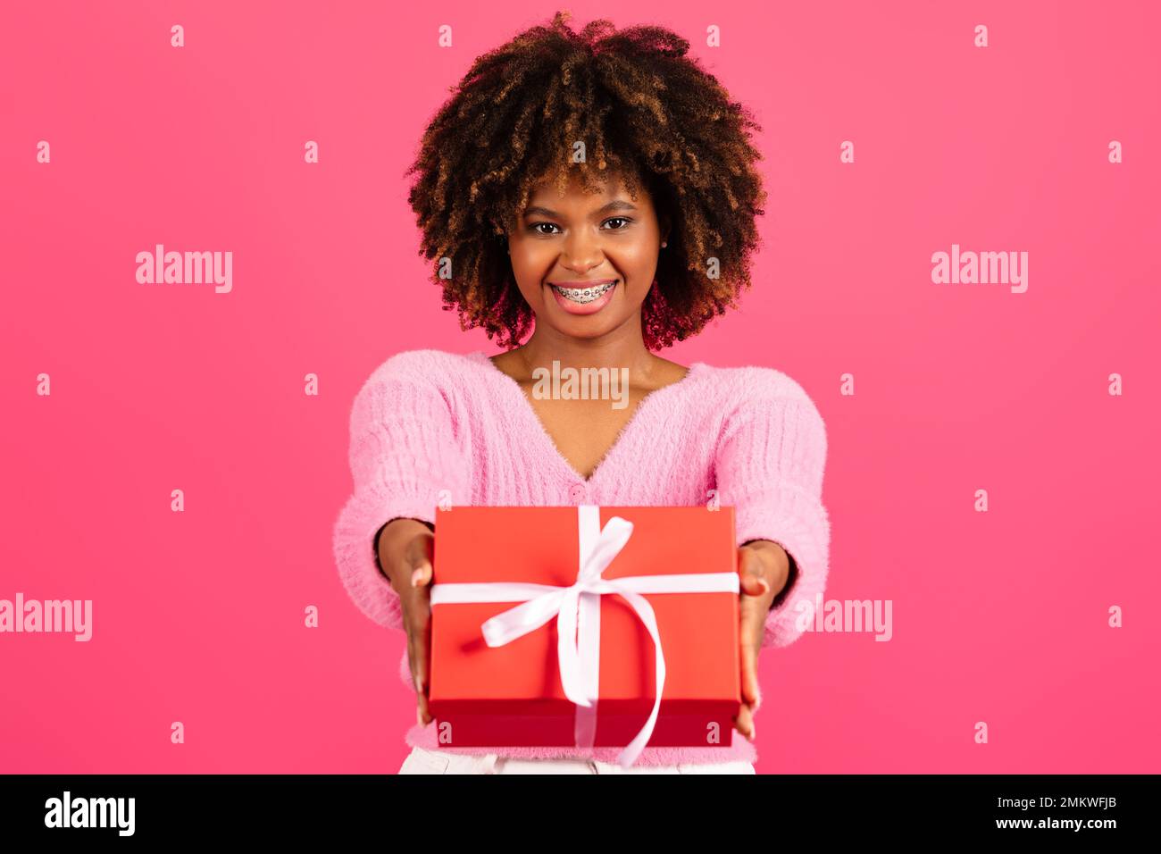 Fröhlich hübsche junge schwarze lockige Frau in legerer Hosenträger gibt rote Box mit Geschenk Stockfoto