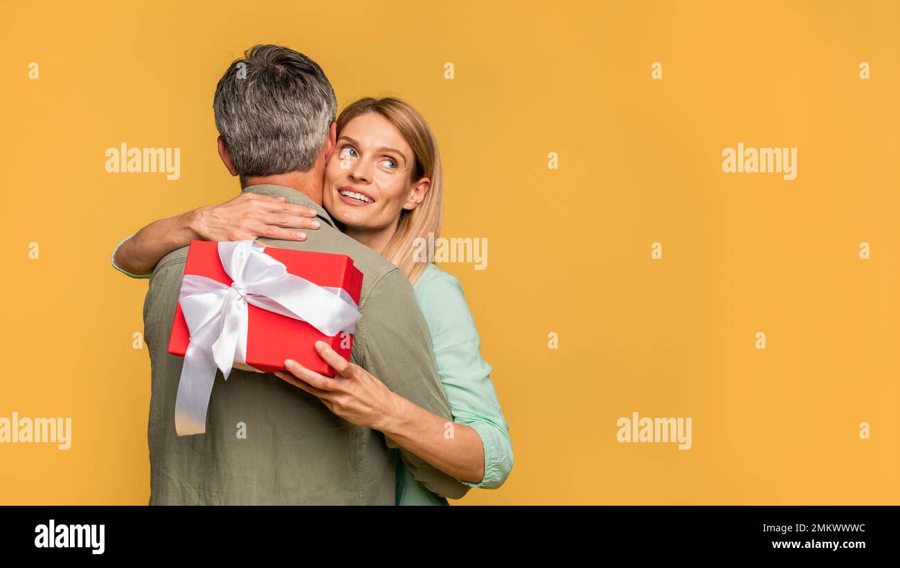 Geschenkkonzept. Liebende Frau mittleren Alters, die ihren Ehemann umarmt und eine Geschenkbox in der Hand hält, die auf die Kopierfläche schaut, Panorama Stockfoto