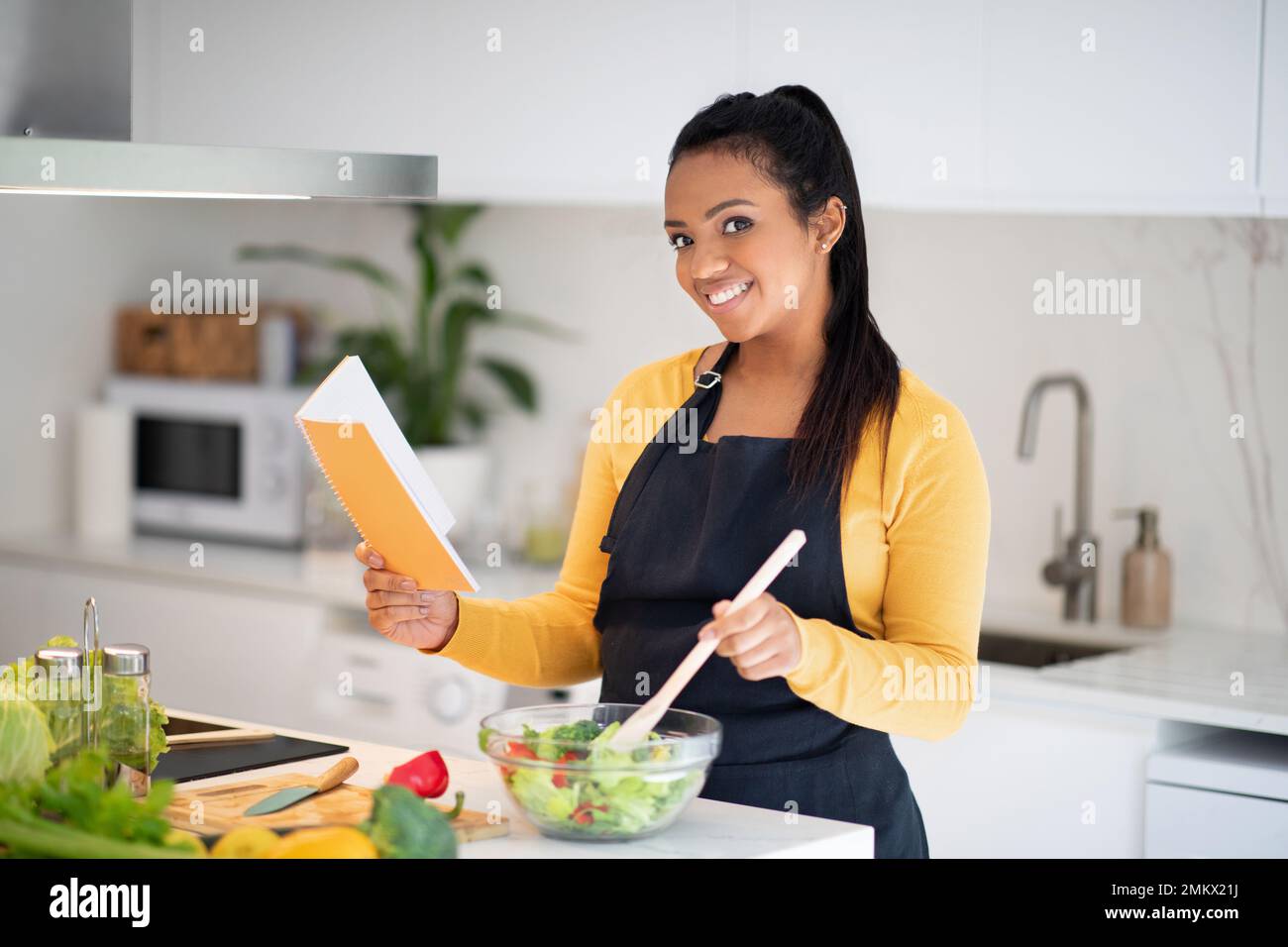 Neues Rezept und hausgemachtes Essen. Die junge afroamerikanische Dame in der Schürze macht Salat, liest Notizbuch Stockfoto