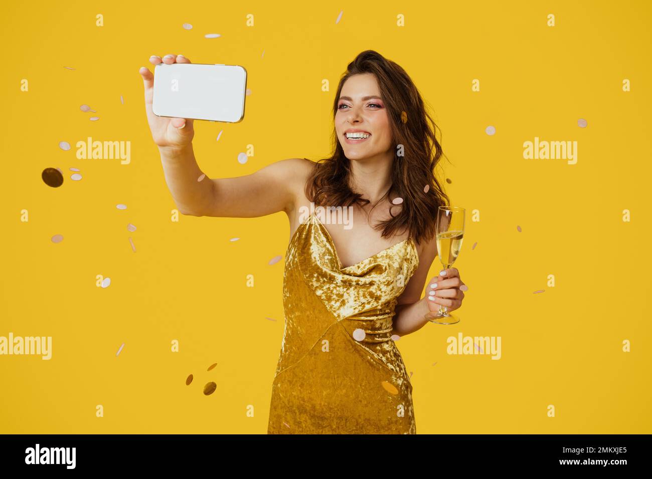 Glückliche Frau in stilvollem Kleid, die Selfie mit einem Glas Champagner macht, gelber Studiohintergrund mit herabfallenden Konfetti Stockfoto