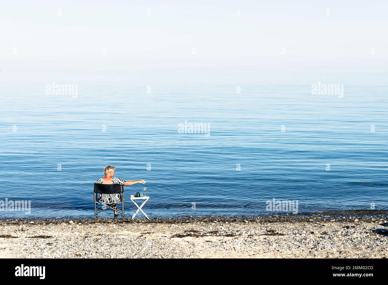 Eine blonde Frau sitzt auf einem Campingstuhl am Ufer von Kattegatt in der Sonne und trinkt Wein, Djursland, Jütland, Dänemark Stockfoto