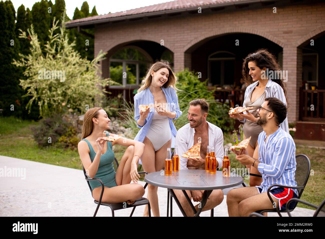 Eine Gruppe von glücklichen jungen Leuten, die mit Apfelwein jubeln und Pizza am Pool im Garten essen Stockfoto